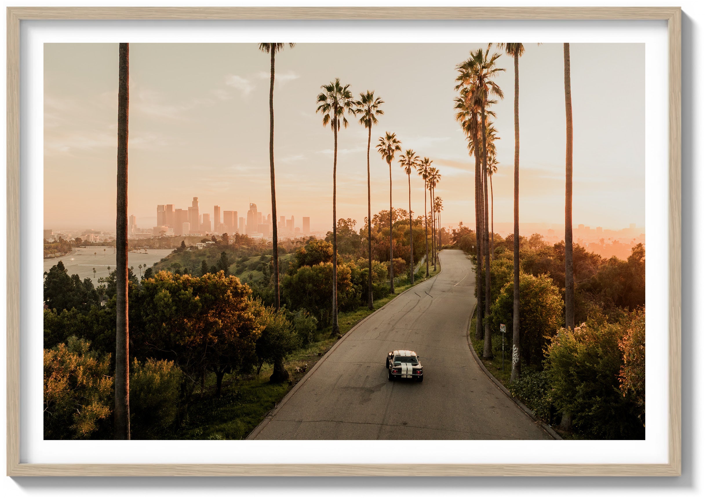 The tall palms rise in clean vertical lines, framing the road as it curves gently through dense, sunlit greenery. The artwork is rendered in color and displayed in a natural wood frame.